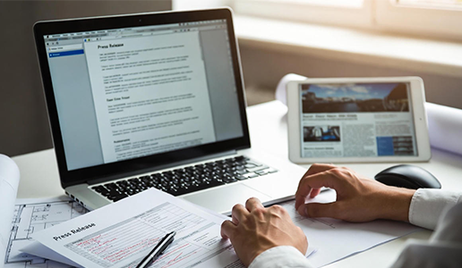 A marketing professional drafting a press release on a laptop and tablet, featuring a physical edited manuscript and a digital news layout.