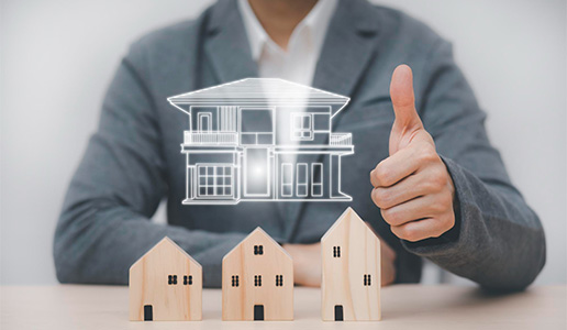 A real estate professional giving a thumbs up behind small wooden house models and a glowing digital blueprint of a luxury home, symbolizing property investment success.